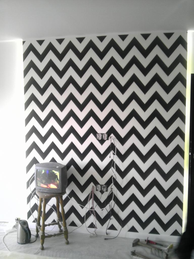 Ściana w zygzaki pomalowana na czarno-biało. Stary telewizor na stołku, widoczne gniazdka elektryczne i narzędzia malarskie na podłodze. Wykończenie wnętrz w trakcie.