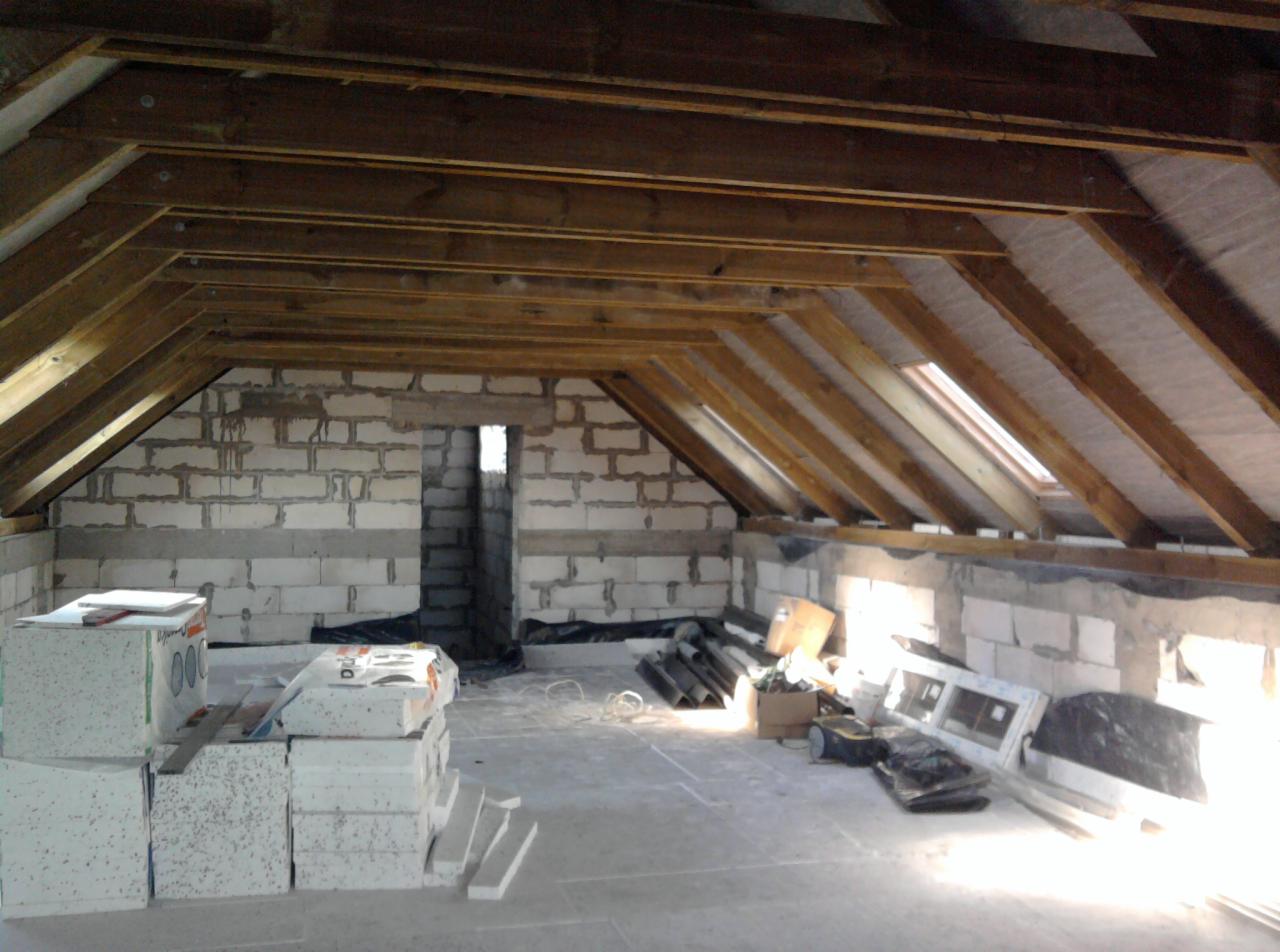 Poddasze w trakcie adaptacji: surowe ściany z betonu komórkowego, drewniana konstrukcja dachu z oknami połaciowymi, materiały budowlane na podłodze. Etap prac wykończeniowych.