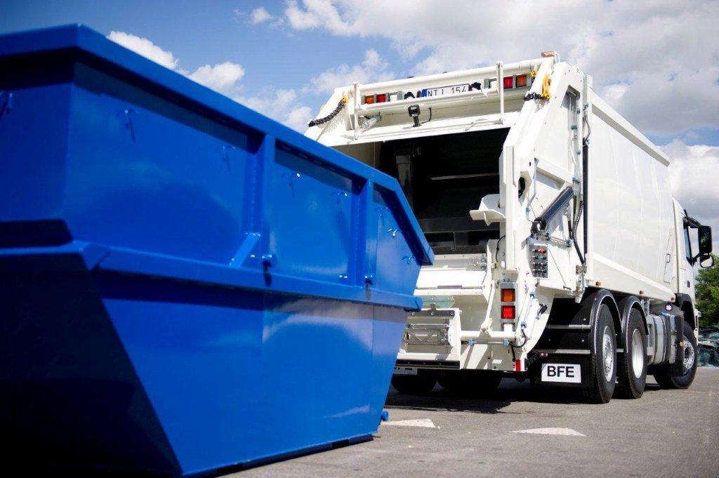Widok z tyłu białej śmieciarki i niebieskiego kontenera na śmieci, z widocznym logo 'BFE' na pojeździe i chmurami na niebie.