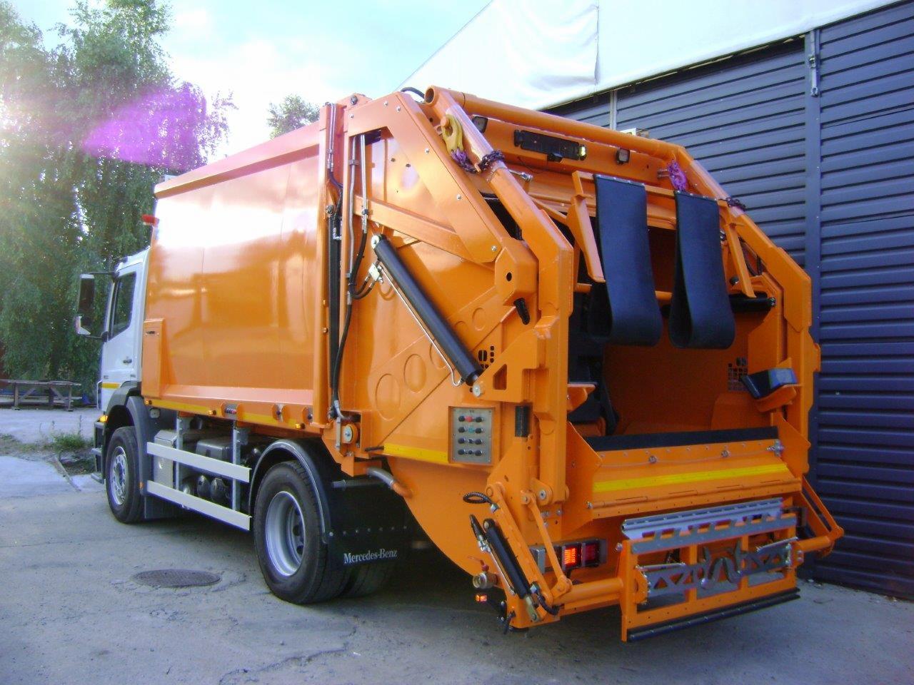 Pomarańczowa śmieciarka Mercedes-Benz BFE Mazur z widocznymi elementami hydraulicznymi i czarnymi gumowymi pasami w tylnej części, zaparkowana na betonowym placu.