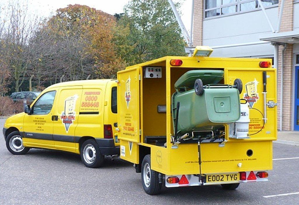 Żółta furgonetka z przyczepą do mycia pojemników na śmieci, z zielonym pojemnikiem odwróconym do góry dnem wewnątrz przyczepy.