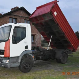 Ciężarówka wywrotka z podniesioną czerwoną skrzynią ładunkową, zaparkowana na trawiastym terenie przed ceglanym domem z anteną satelitarną.