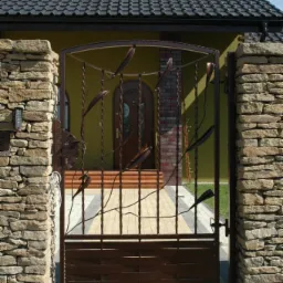 Kuta brama wjazdowa z motywem roślinnym, osadzona między kamiennymi słupkami. W tle widoczny dom z brązowymi elementami i dachem. Brama w kolorze ciemnego brązu.