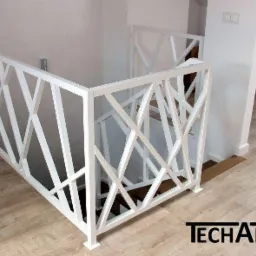 Biała, geometryczna balustrada wewnętrzna na schody, z ukośnymi elementami, widoczna na tle jasnych ścian i podłogi z paneli, z logo firmy w prawym dolnym rogu.