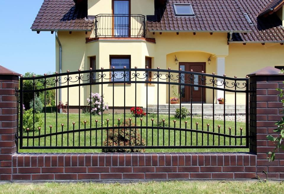 Kute ogrodzenie z czarnych prętów i ceglanymi słupkami na tle żółtego domu z balkonem.