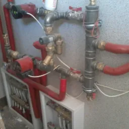 Złożona instalacja z czerwonymi rurami, pompami obiegowymi i rozdzielaczem w białej obudowie na tle ściany o fakturze baranka.