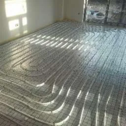 Rozłożone rury ogrzewania podłogowego na siatce w jasnym pomieszczeniu, widoczne okno i folia izolacyjna na ścianie.