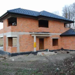 Dwukondygnacyjny dom w stanie surowym otwartym, z ciemną dachówką, widoczne cegły, betonowe nadproża, przygotowane otwory okienne i drzwiowe, teren budowy.