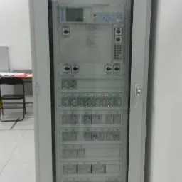 Szara szafa sterownicza z licznymi panelami, przyciskami i wyświetlaczami, widoczna w jasnym pomieszczeniu. Zamknięta szklanymi drzwiami.