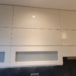 Białe, lakierowane szafki kuchenne wiszące z frontami pełnymi i przeszklonymi, widoczne gniazdka elektryczne w ciemnej ścianie pod szafkami.