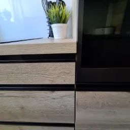 Fragment zabudowy meblowej z drewnianymi frontami szuflad z czarnymi akcentami, na blacie doniczka ze sztuczną trawą i przybory kuchenne.