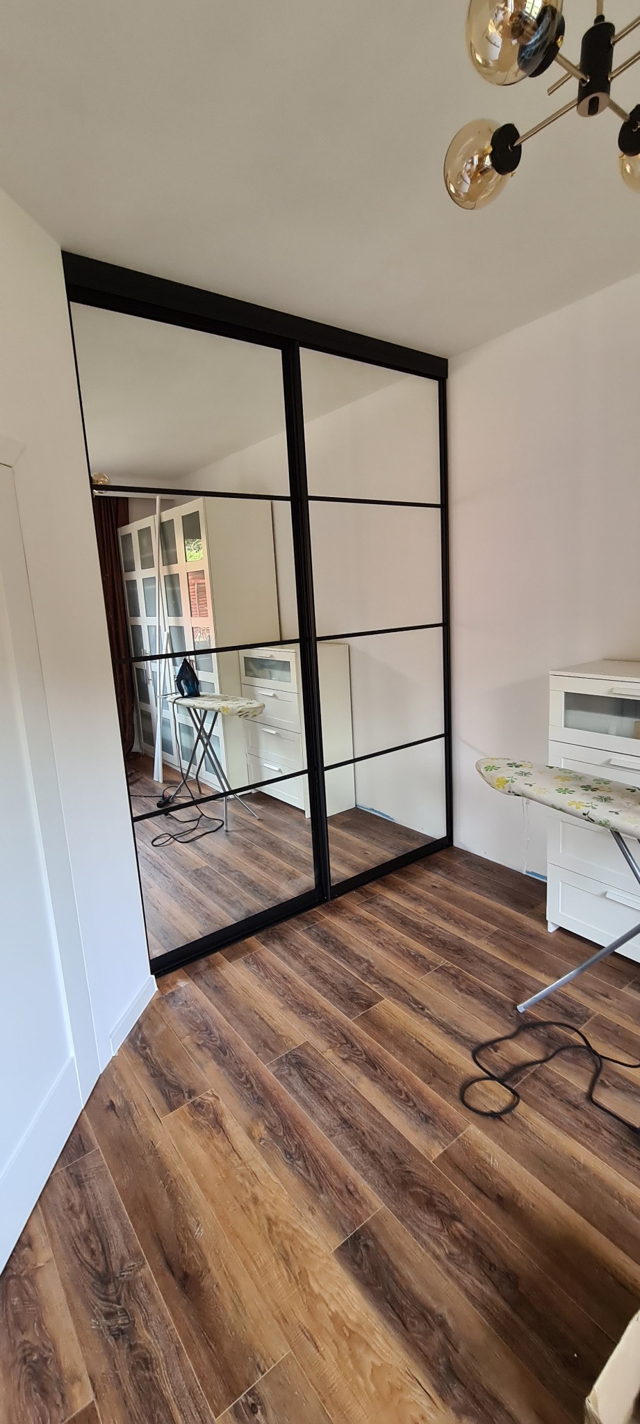Szafa wnękowa z przesuwnymi lustrzanymi drzwiami w czarnej ramie, widoczna na tle drewnianej podłogi, z odbiciem pokoju zawierającym deskę do prasowania i białe meble.