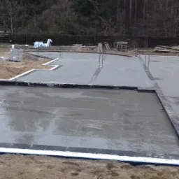 Świeżo wylana, mokra płyta fundamentowa z widocznymi stalowymi prętami zbrojeniowymi i izolacją przeciwwilgociową, otoczona piaskiem, z zaparkowanym białym vanem w tle.