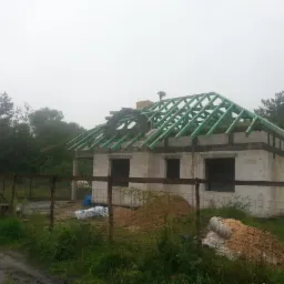 Surowy stan domu z bloczków betonowych w trakcie budowy dachu, widoczne zielone krokwie i komin, teren ogrodzony siatką, pochmurna pogoda.