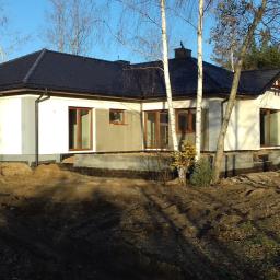 Usługi Ogólno-Budowlane - Niedokończony, parterowy dom z ciemnym dachem na tle lasu, widoczne fundamenty i częściowo otynkowana elewacja w kolorze białym.