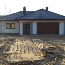 Parterowy dom jednorodzinny z brązową bramą garażową i ciemną dachówką, otoczony piaszczystym terenem z widocznymi śladami opon i niebieską rurą kablową, w tle drzewa bez liści.