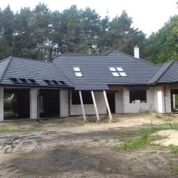 Usługi Ogólno-Budowlane - Niedokończony dom murowany z czarną dachówką i widocznymi lukarnami, na tle lasu, z podporami z drewna i stertą piasku na pierwszym planie.
