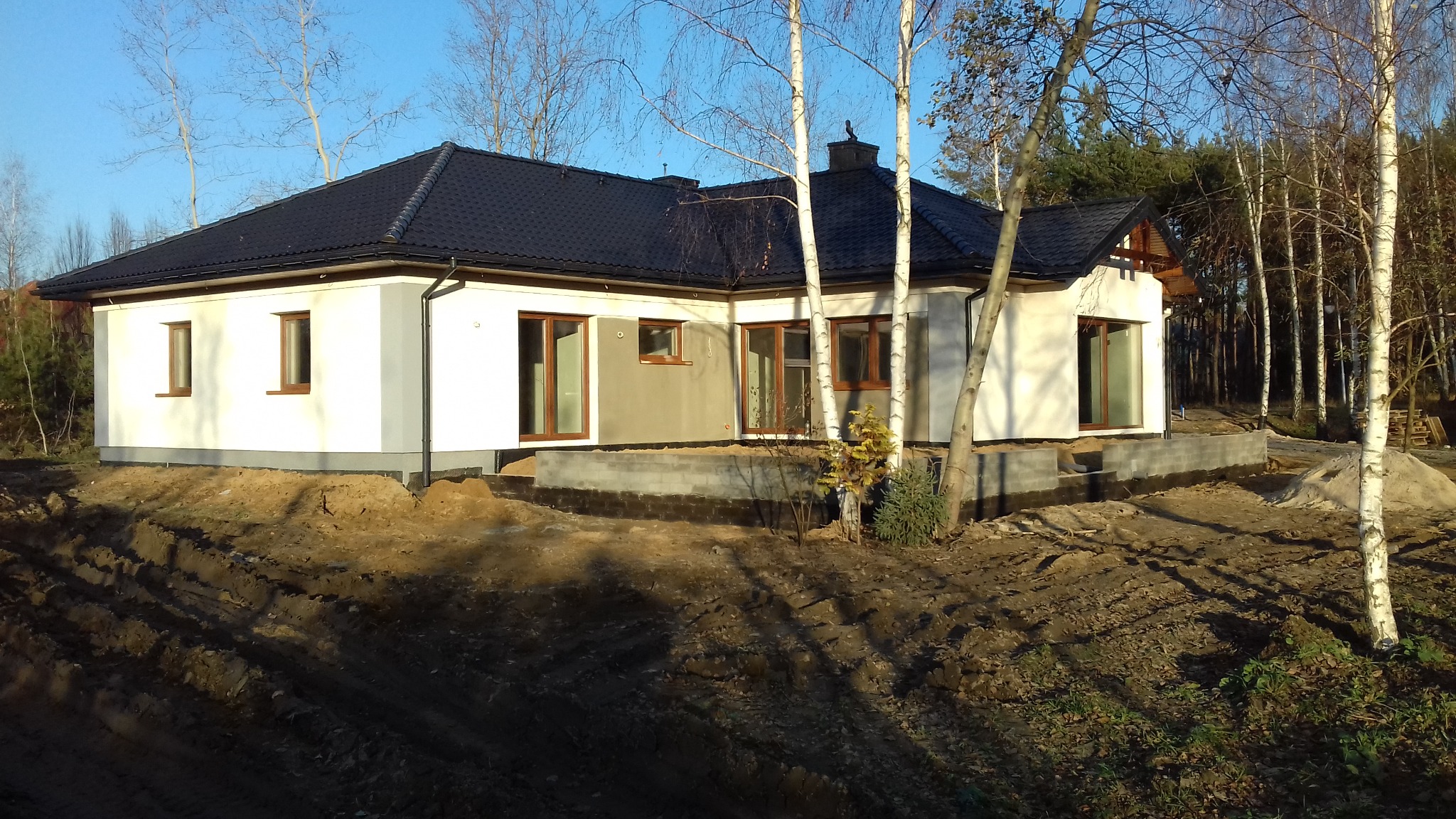 Niedokończony, parterowy dom z ciemnym dachem na tle lasu, widoczne fundamenty i częściowo otynkowana elewacja w kolorze białym.