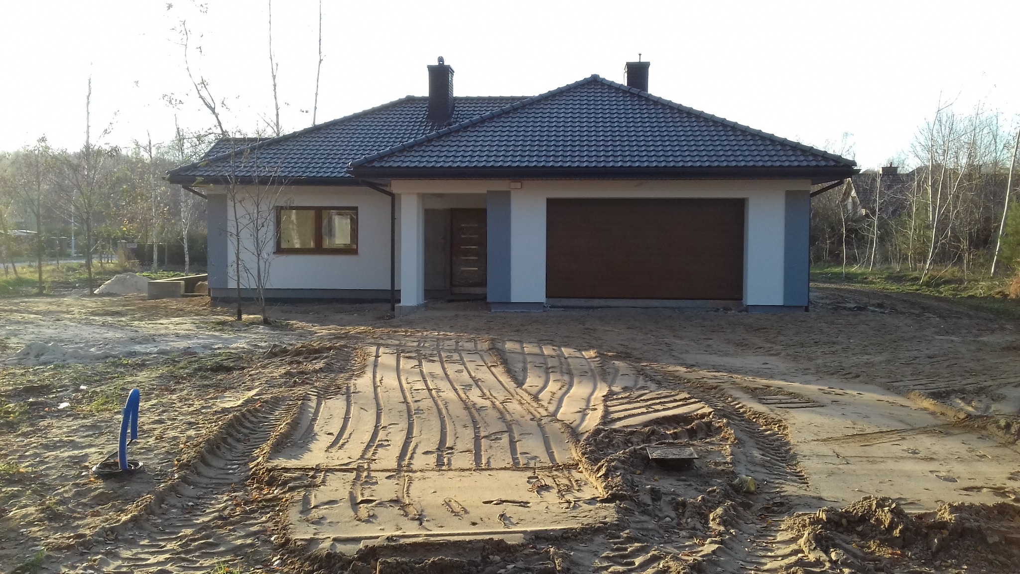 Parterowy dom jednorodzinny z brązową bramą garażową i ciemną dachówką, otoczony piaszczystym terenem z widocznymi śladami opon i niebieską rurą kablową, w tle drzewa bez liści.
