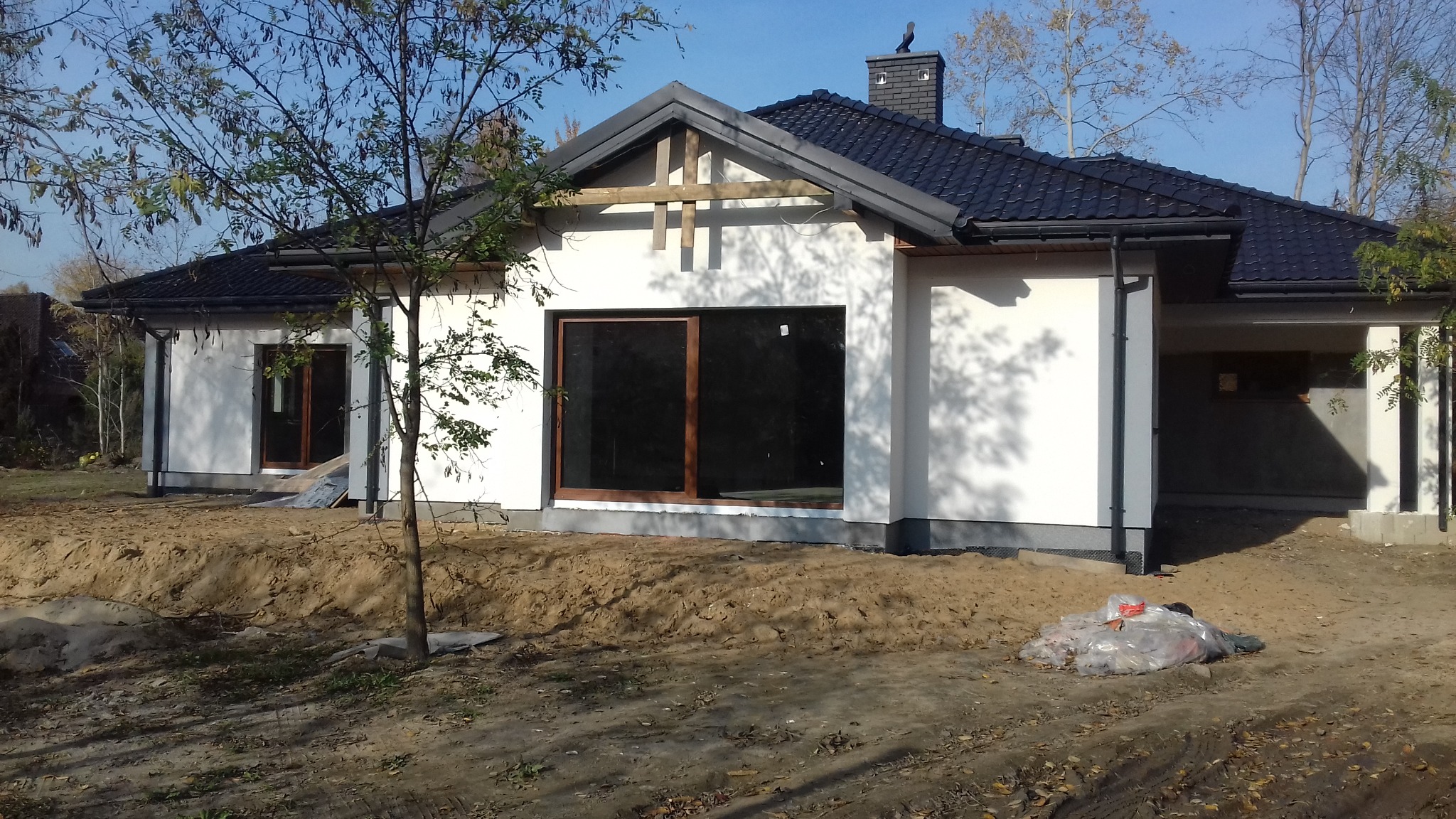 Niedokończony parterowy budynek z ciemną dachówką, dużym oknem i elementami drewnianymi nad nim, otoczony ziemią i młodym drzewem.