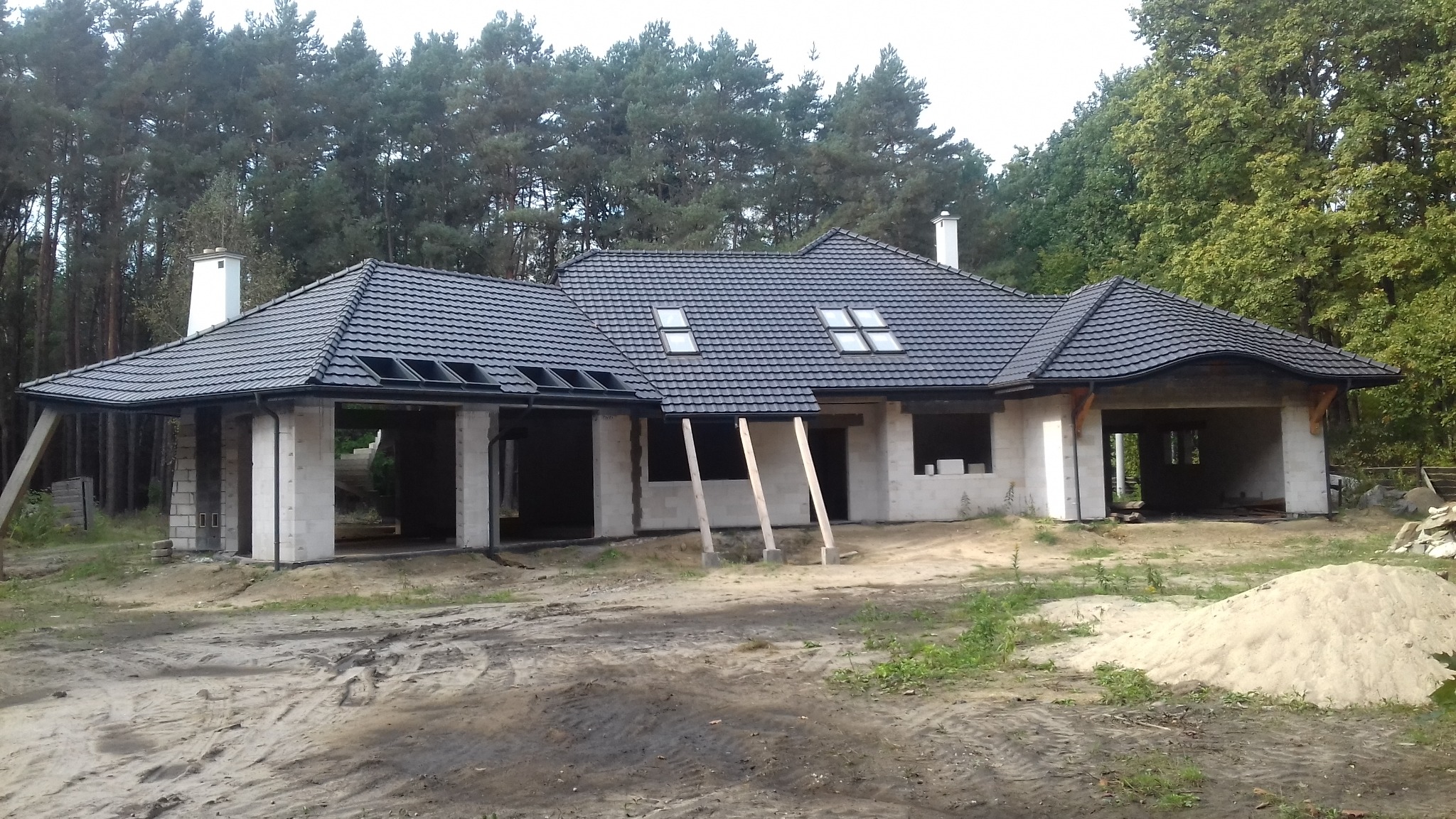 Niedokończony dom murowany z czarną dachówką i widocznymi lukarnami, na tle lasu, z podporami z drewna i stertą piasku na pierwszym planie.