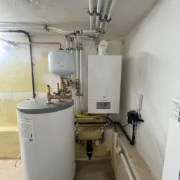 Bojler z zasobnikiem i piec gazowy De Dietrich zamontowane w pomieszczeniu z surowymi ścianami i instalacjami pod sufitem.