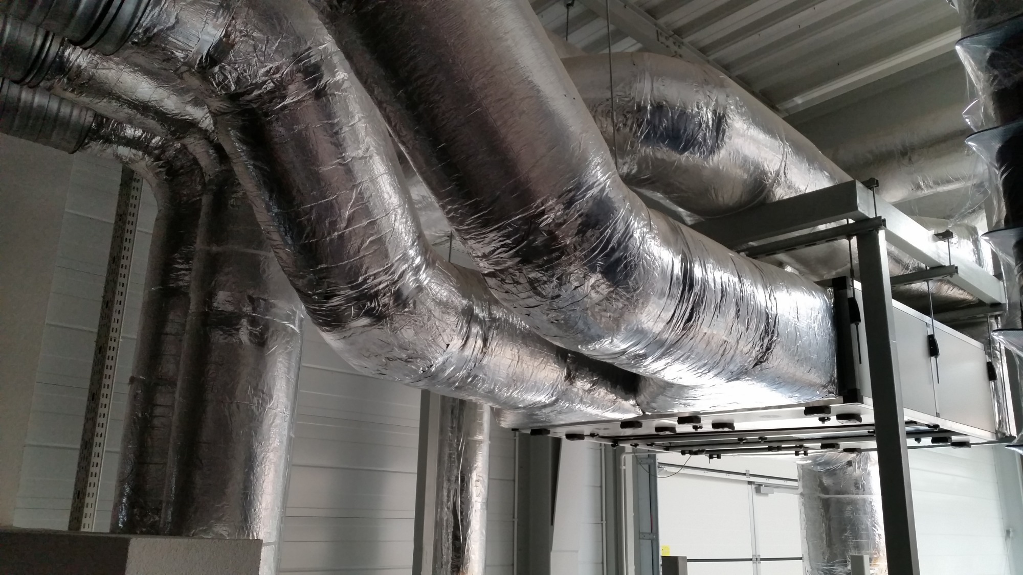System kanałów wentylacyjnych owiniętych folią izolacyjną, zamontowany pod sufitem w hali przemysłowej, widoczne elementy konstrukcji wsporczej i fragment białej ściany z blachy trapezowej.
