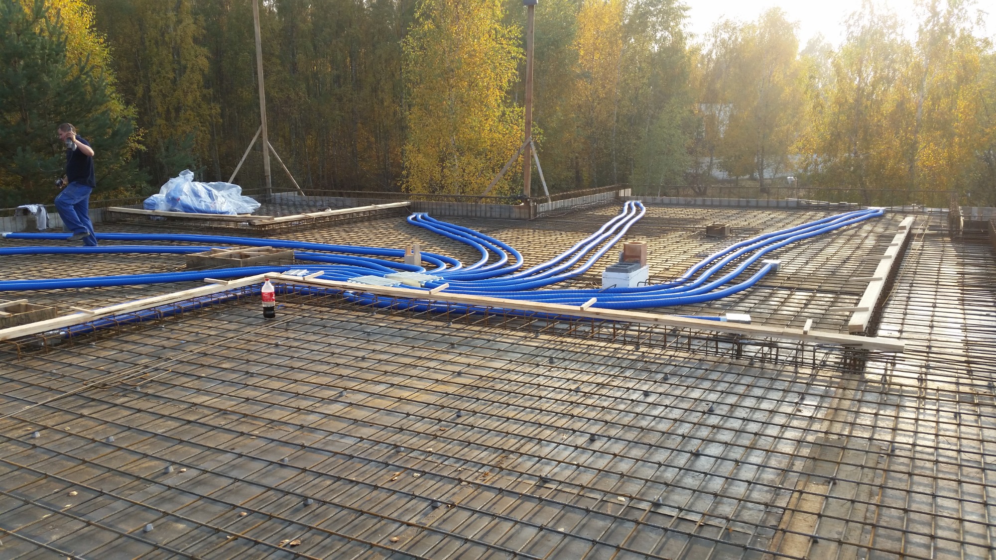 Niebieskie rury wentylacyjne ułożone na zbrojeniu stropu, widoczny pracownik w tle i butelka napoju przy krawędzi, jesienne drzewa w tle.
