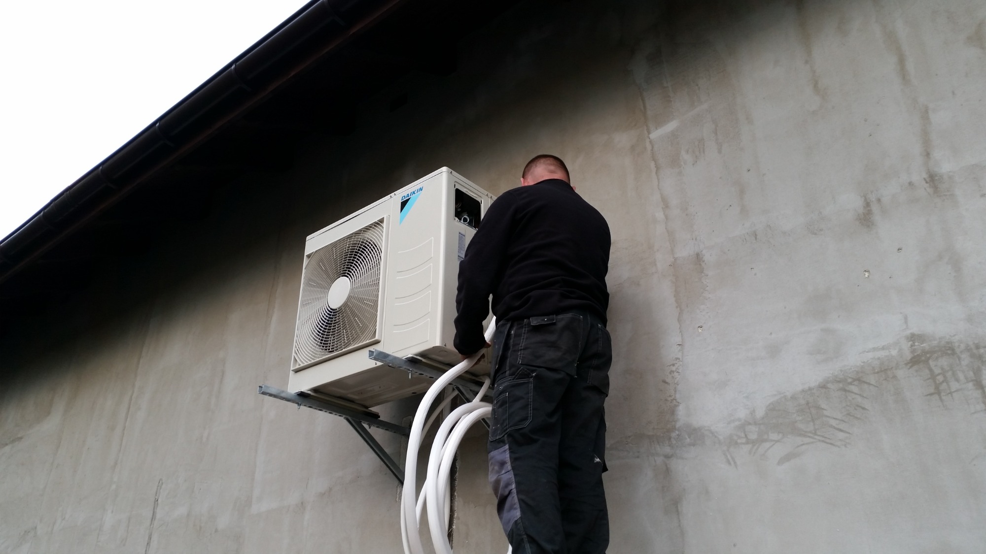 Instalacja zewnętrznej jednostki klimatyzacji Daikin na szarej ścianie budynku; widoczne liczne białe przewody i monter w czarnym ubraniu na drabinie.