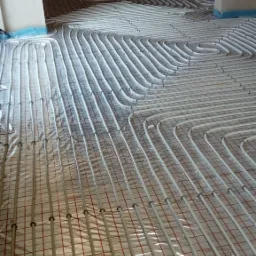 Białe rurki ogrzewania podłogowego ułożone na folii z czerwoną siatką w surowym wnętrzu, widok z góry pod kątem, przygotowane do zalania wylewką.