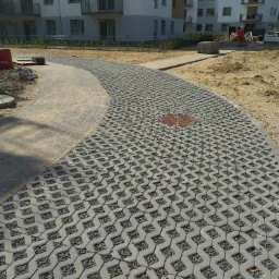 Ścieżka z ażurowych płyt betonowych wypełnionych grysem, prowadząca przez piaszczysty teren budowy w kierunku nowych budynków mieszkalnych, z widoczną studzienką kanalizacyjną.