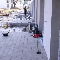 Szare prostokątne płyty chodnikowe ułożone w rzędach, z pracownikiem w żółtym kasku w tle i narzędziami budowlanymi w pobliżu ściany budynku.