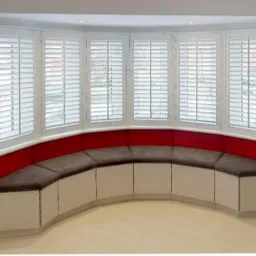 Wnęka okienna z białymi drewnianymi żaluzjami i wbudowanym siedziskiem z czerwonym oparciem i brązowymi poduszkami, Krotoszyny Pomorskie.