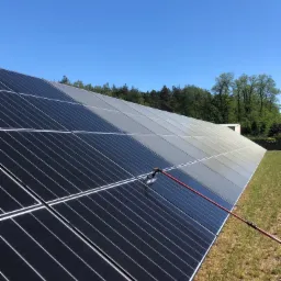 Panel słoneczny czyszczony szczotką na długim wysięgniku, widoczna różnica w czystości pomiędzy poszczególnymi panelami, na tle błękitne niebo i zieleń drzew.