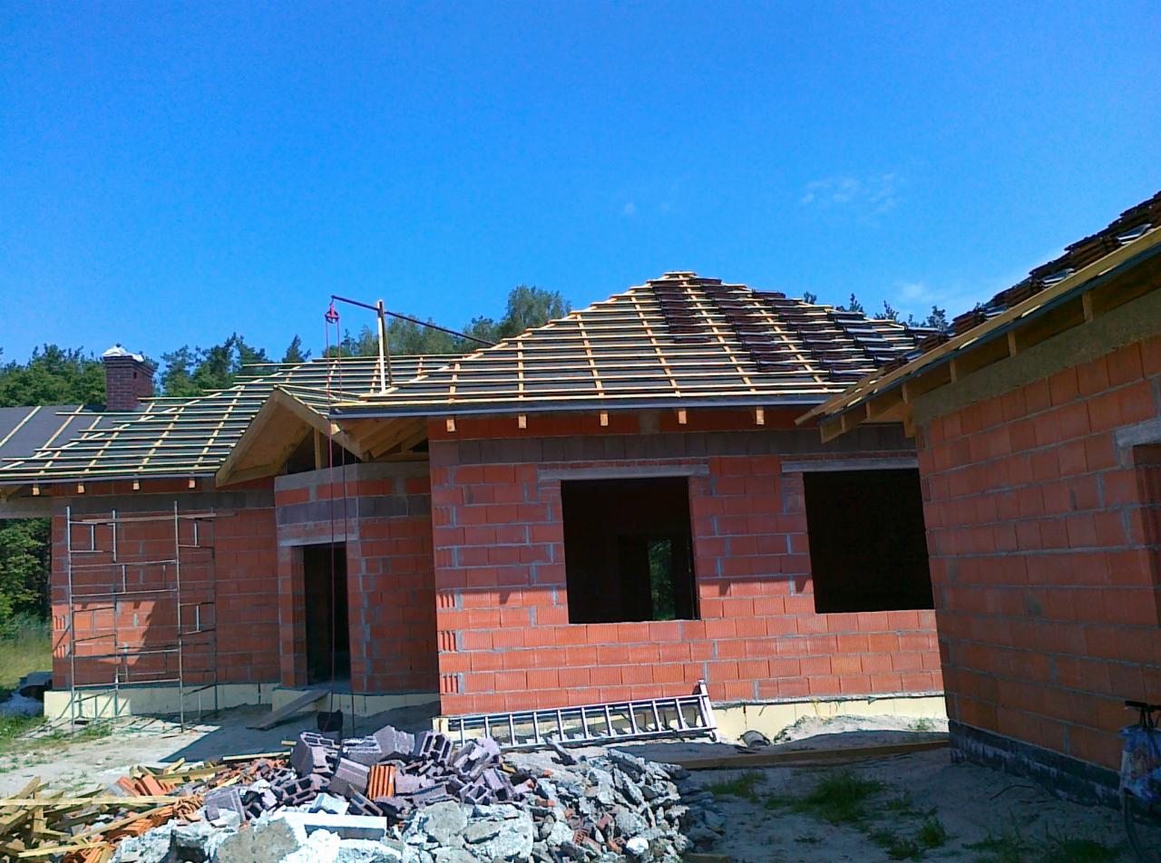 Budowa domu murowanego z czerwonej cegły: wzniesione ściany, częściowo ułożony dach z łatami, na ziemi gruz i drabina, w tle las i błękitne niebo.
