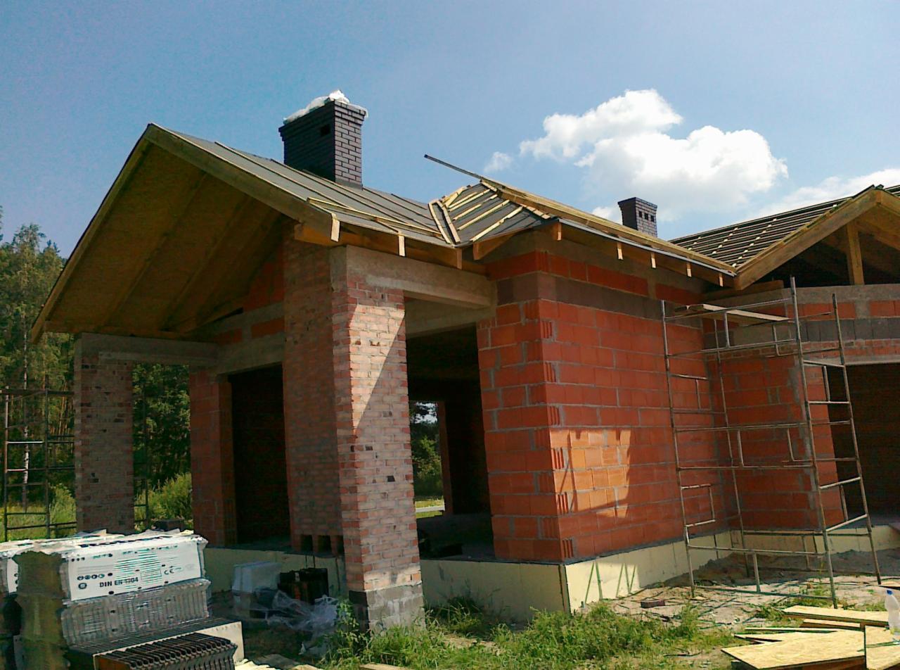 Surowy budynek jednorodzinny z czerwonej cegły w trakcie budowy, widoczne rusztowanie, elementy więźby dachowej i komin, na pierwszym planie materiały budowlane.