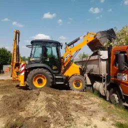 Żółto-czarna koparko-ładowarka JCB załadowuje ziemię do pomarańczowej wywrotki Volvo na placu budowy w słoneczny dzień.