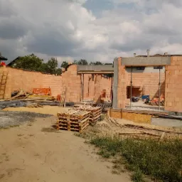 Wznoszenie ścian budynku z cegły ceramicznej Heluz, widoczne palety z materiałem, częściowo wylane fundamenty i elementy szalunków, budowa w trakcie realizacji.