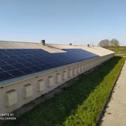 Długi budynek gospodarczy z szarym dachem pokrytym panelami słonecznymi. Widoczny fragment drogi z kostki, trawnik, oraz turbina wiatrowa w oddali.