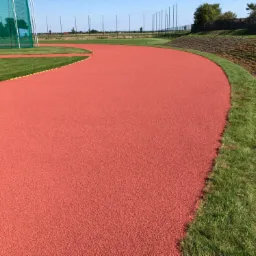 Czerwona, syntetyczna bieżnia lekkoatletyczna zakręca łagodnym łukiem, otoczona pasem zielonej trawy i nasypem ziemnym obsadzonym roślinnością.