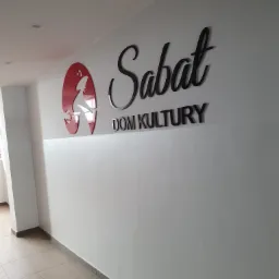 Logo 'Sabat Dom Kultury' z podświetlanym czerwonym elementem graficznym na białej ścianie w korytarzu, czarne litery w różnych stylach.