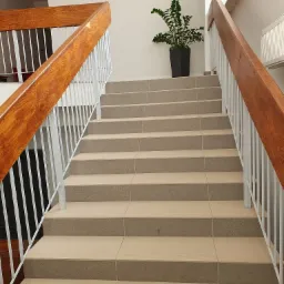 Schody wyłożone płytkami ceramicznymi w budynku biurowym, z drewnianą balustradą i rośliną doniczkową na górnym stopniu.