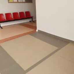 Beżowa podłoga wyłożona płytkami w biurze z szarymi akcentami przy ścianie i w pasach pomiędzy płytkami, w tle rząd czerwonych krzeseł i tablica ogłoszeń.