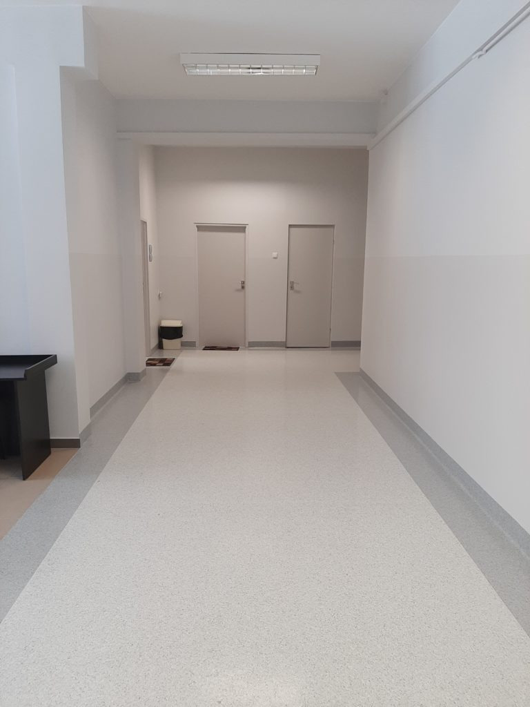 Długi korytarz w budynku biurowym z jasną, nakrapianą wykładziną podłogową i szarą listwą przypodłogową, dwie białe drzwi na końcu korytarza, widoczny fragment czarnego biurka z lewej strony.