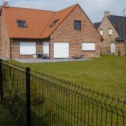 Nowoczesny dom jednorodzinny z czerwonej cegły z tarasem i roletami zewnętrznymi, ogrodzony czarnym metalowym płotem panelowym.