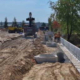 KERBUD - Budowa drogi: koparka Volvo, wykop, betonowe elementy kanalizacji deszczowej, pracownicy w kaskach i kamizelkach odblaskowych, drzewa w tle.