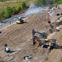 KERBUD - Szeroki kadr z góry: żółto-czarna koparka Volvo w akcji na placu budowy drogi. W tle walec, pracownicy w kaskach i betonowe elementy. Dym unoszący się w powietrzu.