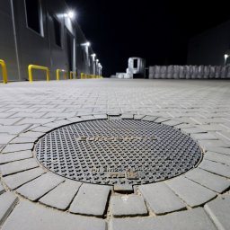 KERBUD - Szara kostka brukowa z okrągłą studzienką kanalizacyjną na pierwszym planie, oświetlona lampami ulicznymi w Katowicach. W tle budynek magazynowy.