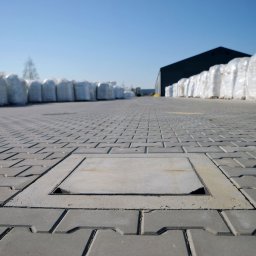 KERBUD - Szara kostka brukowa ułożona na placu, z włazem rewizyjnym pośrodku. W tle rząd worków z materiałem i budynek magazynowy.