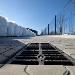 KERBUD - Szara kostka brukowa z kratką ściekową w centrum kadru, w tle rząd worków i niebieskie ogrodzenie panelowe. Ujęcie z dołu, podkreślające perspektywę.
